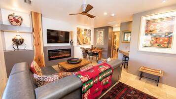 A modern living room with a gray sofa, a wooden coffee table, and a red blanket. There is a fireplace, a ceiling fan, and framed artwork on the walls.