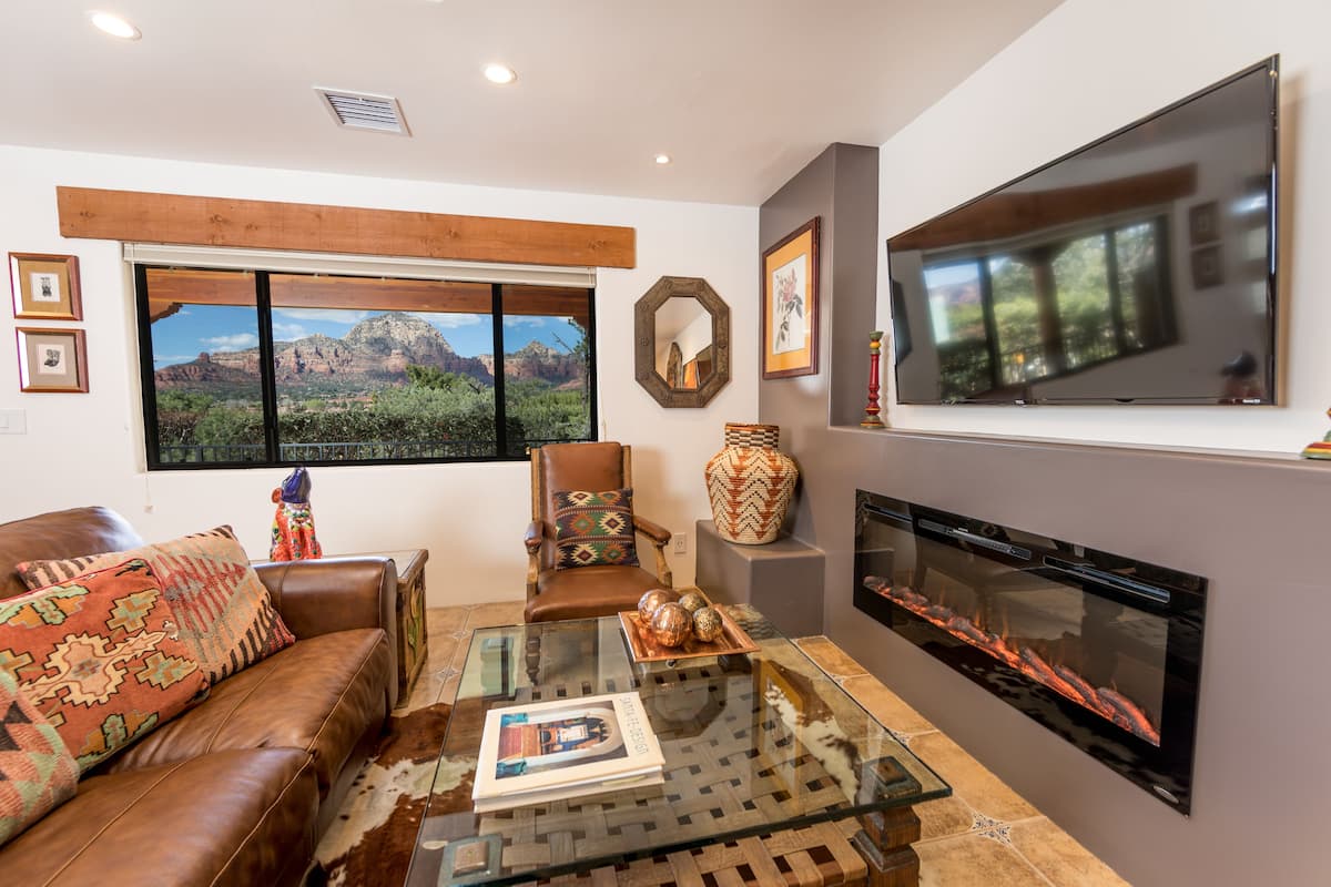 A living room with a leather sofa, a glass coffee table, a fireplace, and a large window with a mountain view.