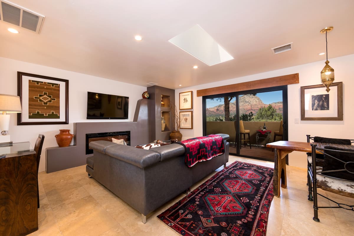 A modern living room with a gray sofa, a fireplace, a dining area with a table and chairs, and a large window with a mountain view.