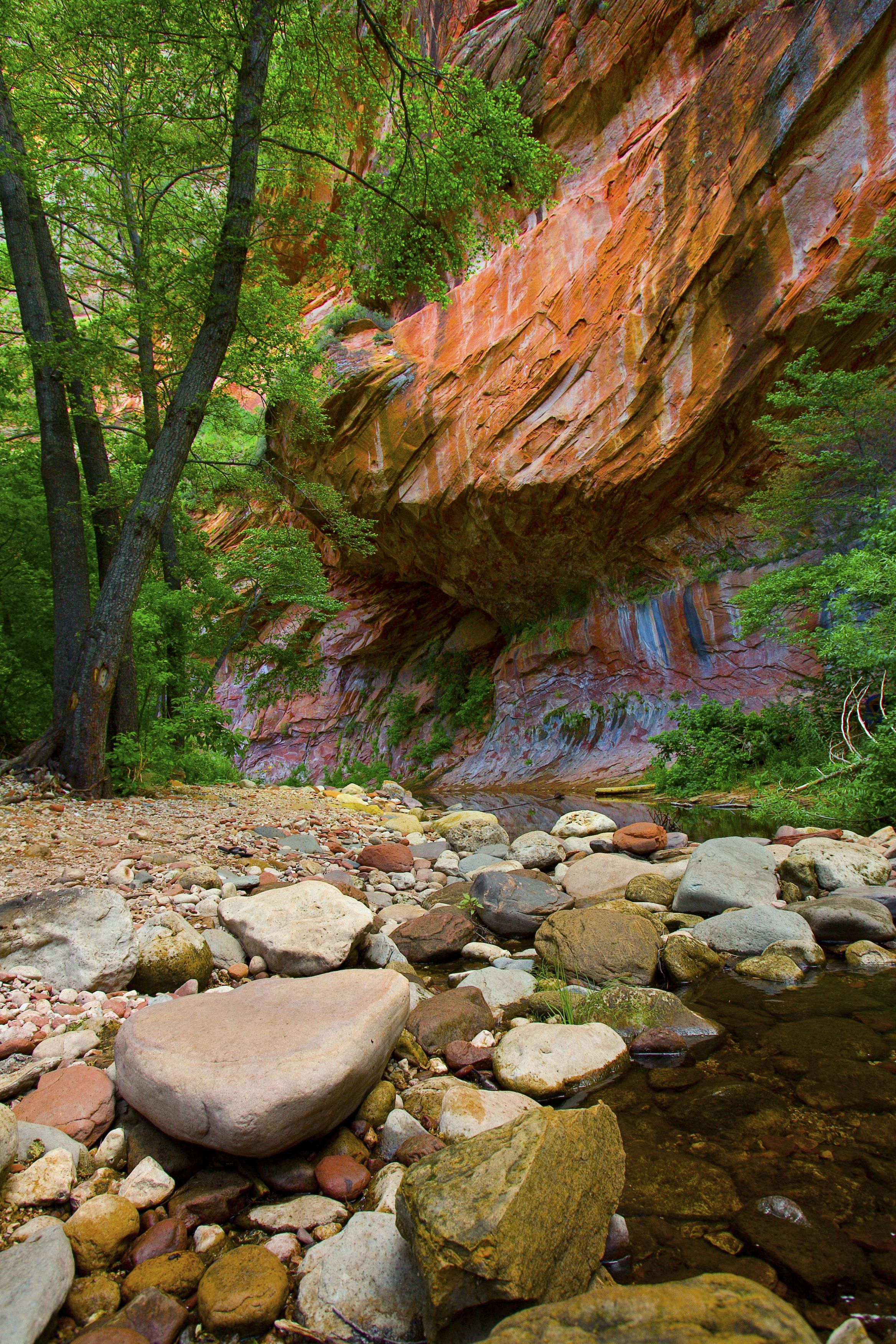 A natural landscape with a rocky streambed, trees, and a large red rock formation.