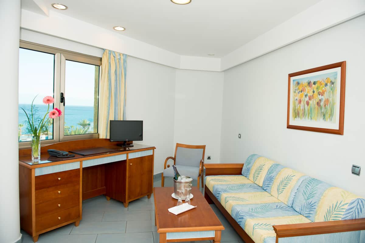 A room with a wooden desk, a television, a sofa, and a window with a view of the sea.