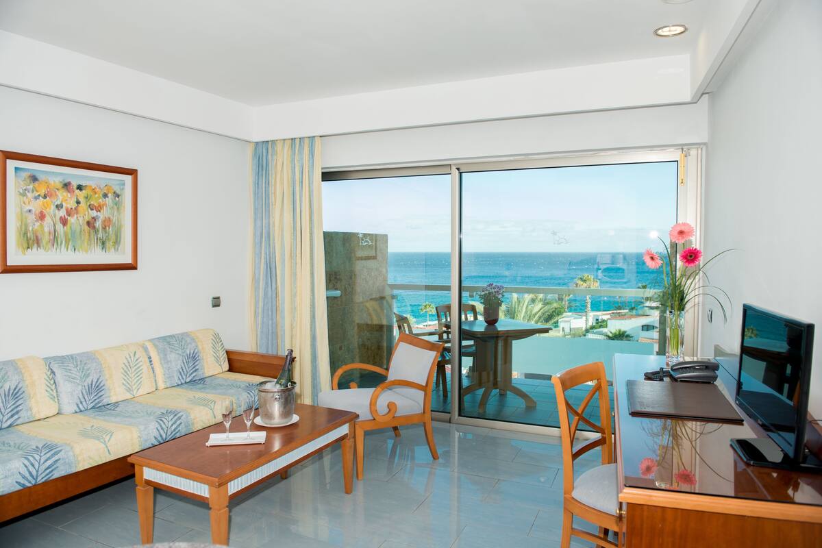 A modern hotel room with a balcony, a sofa, a dining table, and a TV.