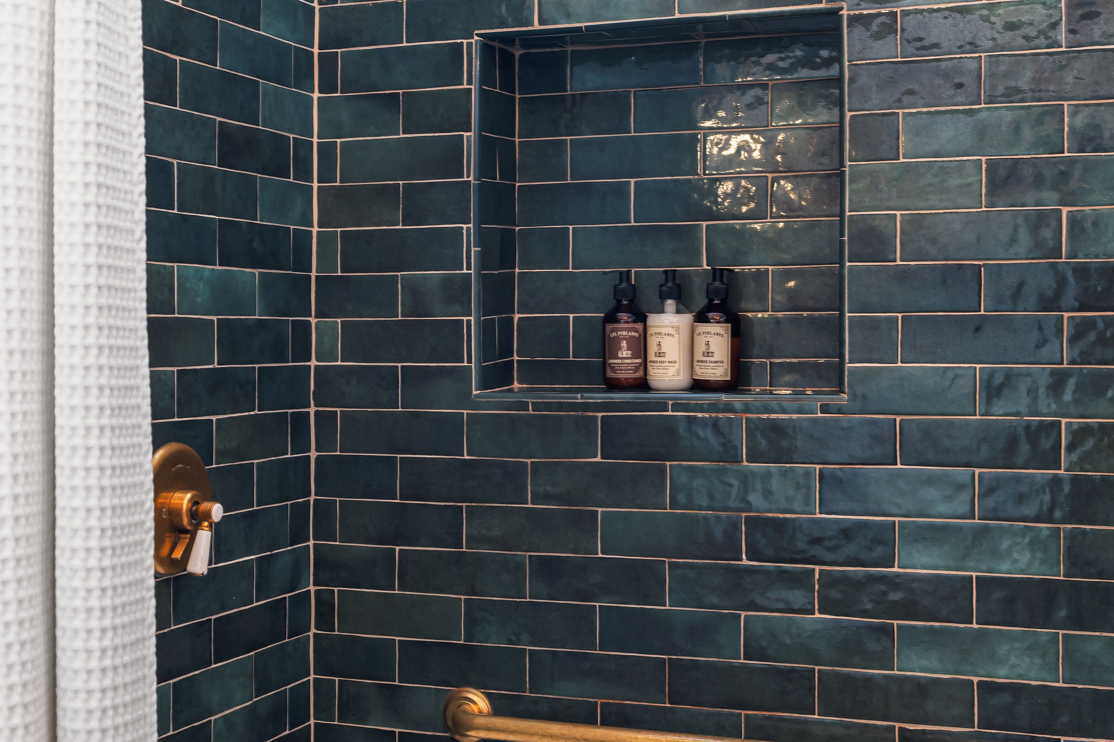A bathroom with dark blue tiled walls, a shower area, and a shelf with three bottles of toiletries.