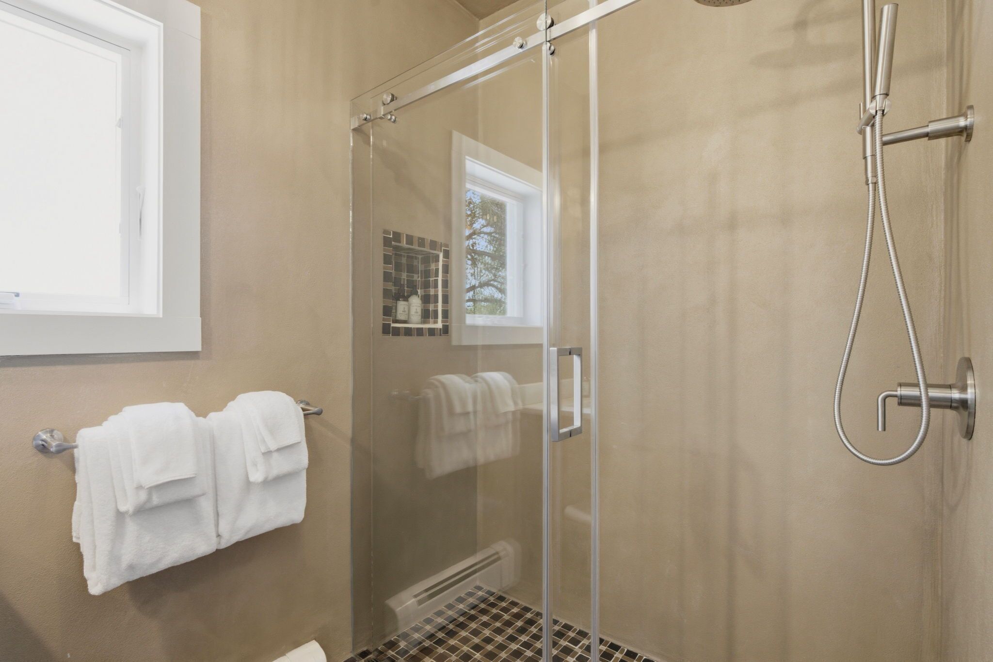 Une douche fermée par des parois en verre, dotée d’un pommeau de douche à main, d’un porte-serviettes avec des serviettes et d’une fenêtre donnant sur les arbres.