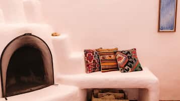 A white fireplace with a screen, a small basket below it, and three colorful pillows on a white shelf against a plain wall.