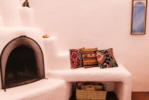 A white fireplace with a screen, a small basket below it, and three colorful pillows on a white shelf against a plain wall.