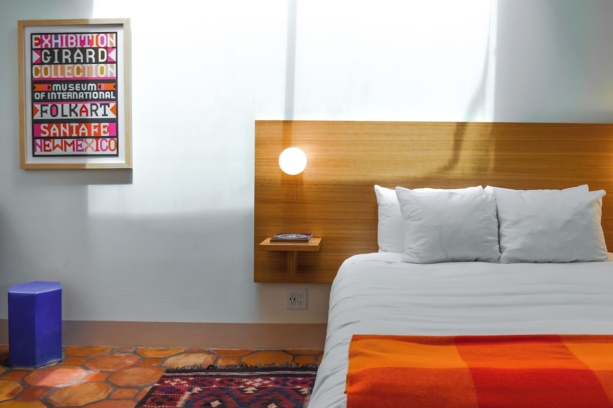 Une chambre d’hôtel avec une tête de lit en bois, deux lampes de chevet, un lit avec du linge de lit blanc, un tapis coloré et un panneau décoratif accroché au mur.