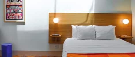 Habitación de hotel con cabecera de madera, dos lámparas de noche, cama con ropa blanca, alfombra colorida y un cartel enmarcado en la pared.
