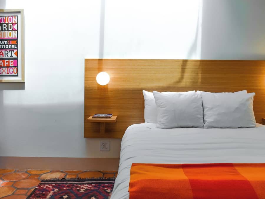 Quarto de hotel com cabeceira de madeira, duas luminárias de apoio, cama com roupa de cama branca, tapete colorido e um letreiro emoldurado na parede.