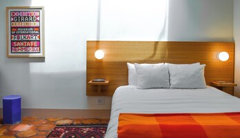 A hotel room with a wooden headboard, two bedside lamps, a bed with white linens, a colorful rug, and a framed sign on the wall.