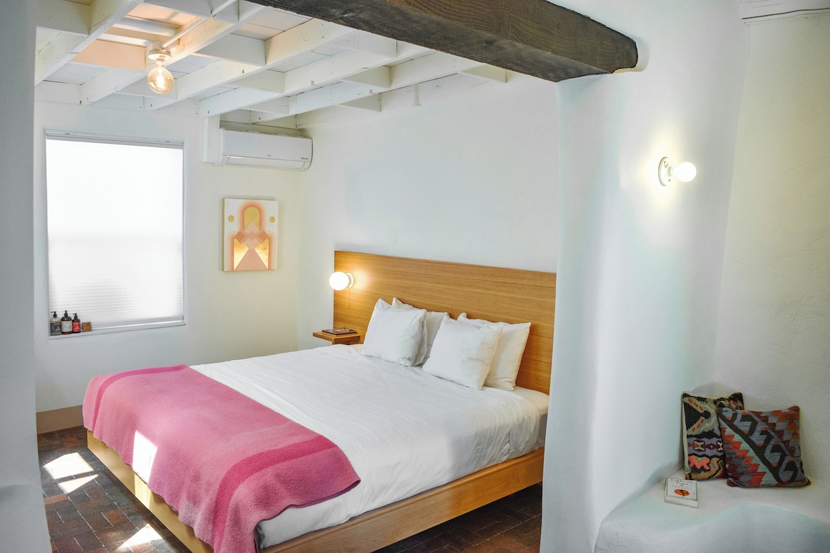 Une chambre à coucher avec un lit en bois, une literie blanche, une couverture rose, une table de chevet avec une lampe, une fenêtre avec des stores et un climatiseur fixé au mur.