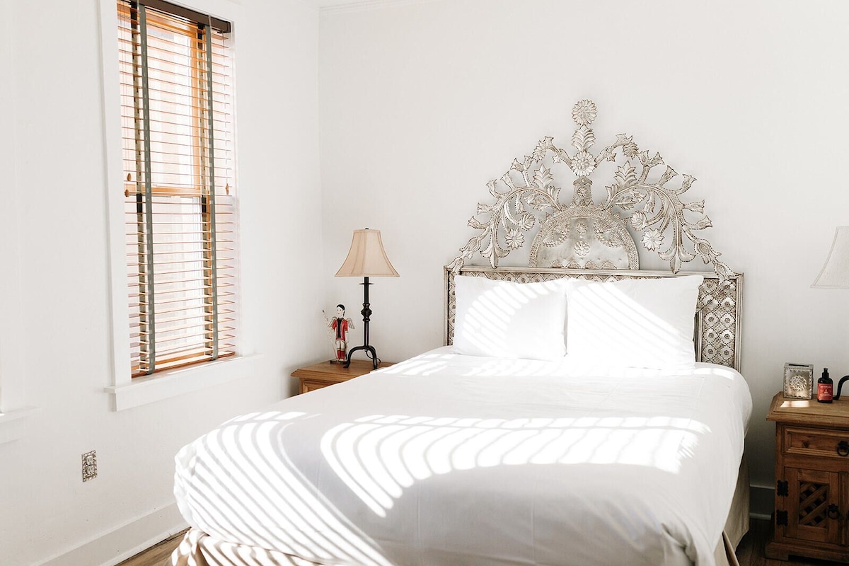 Une chambre à coucher avec un lit blanc, une tête de lit décorative, une lampe de chevet et une fenêtre avec des stores.
