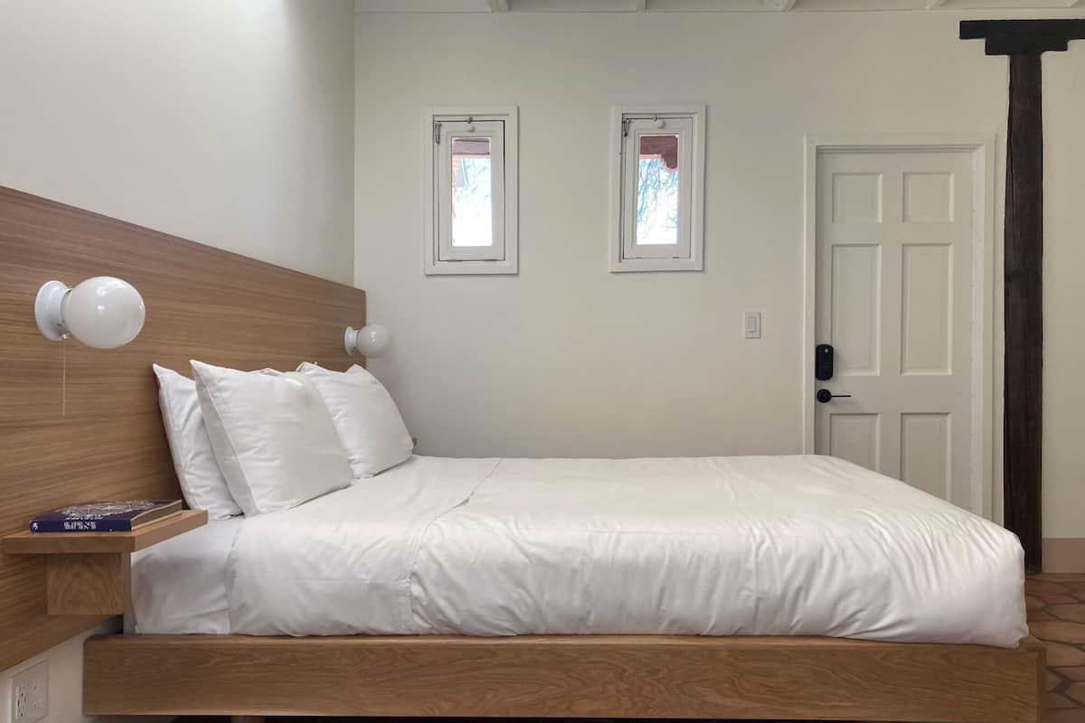 Une chambre à coucher avec un cadre de lit en bois, du linge de lit blanc, deux tables de chevet avec des lampes et deux fenêtres avec des rideaux.