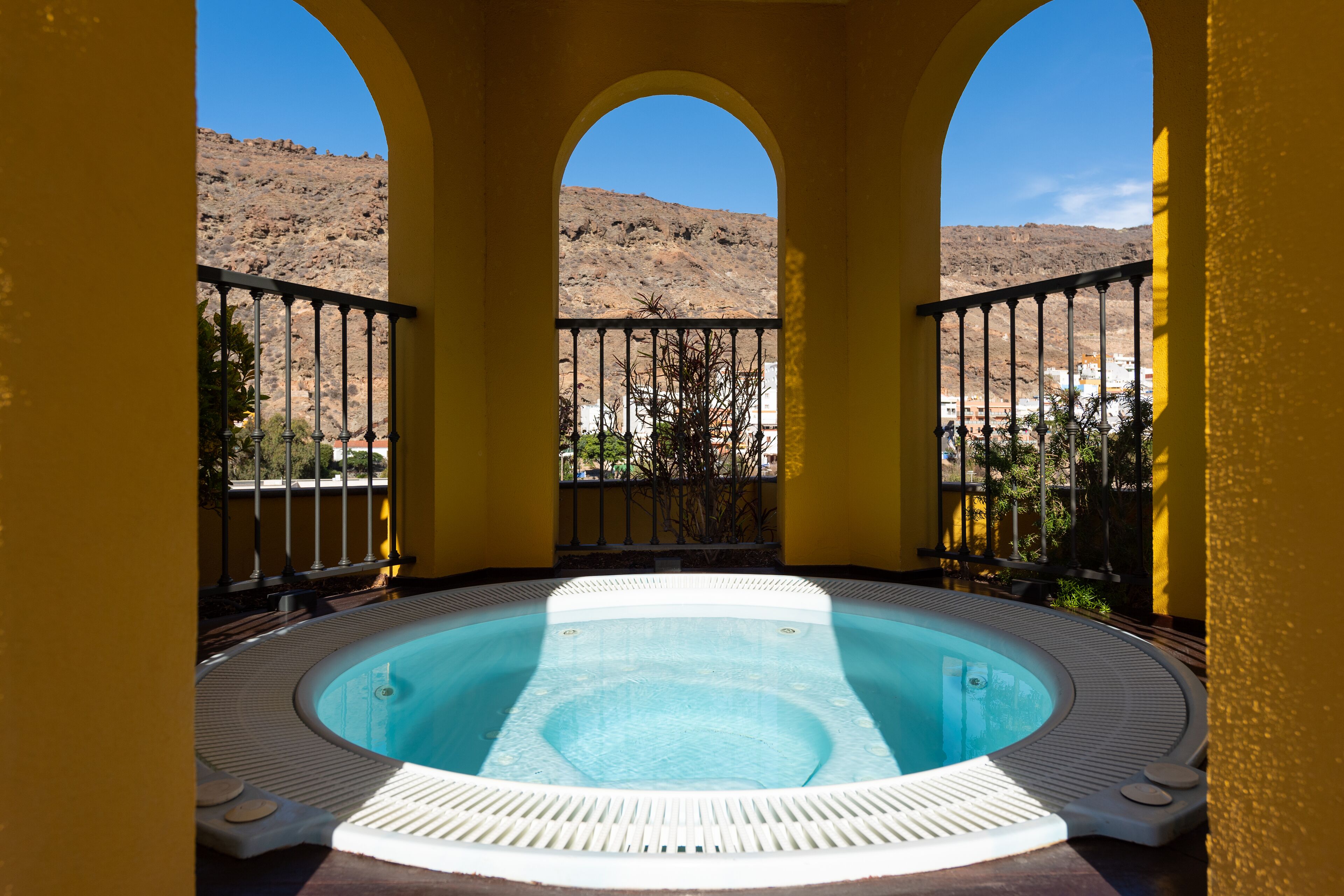 Un bain à remous offrant une vue sur un paysage montagneux, avec en arrière-plan un bâtiment jaune doté d’arcades.