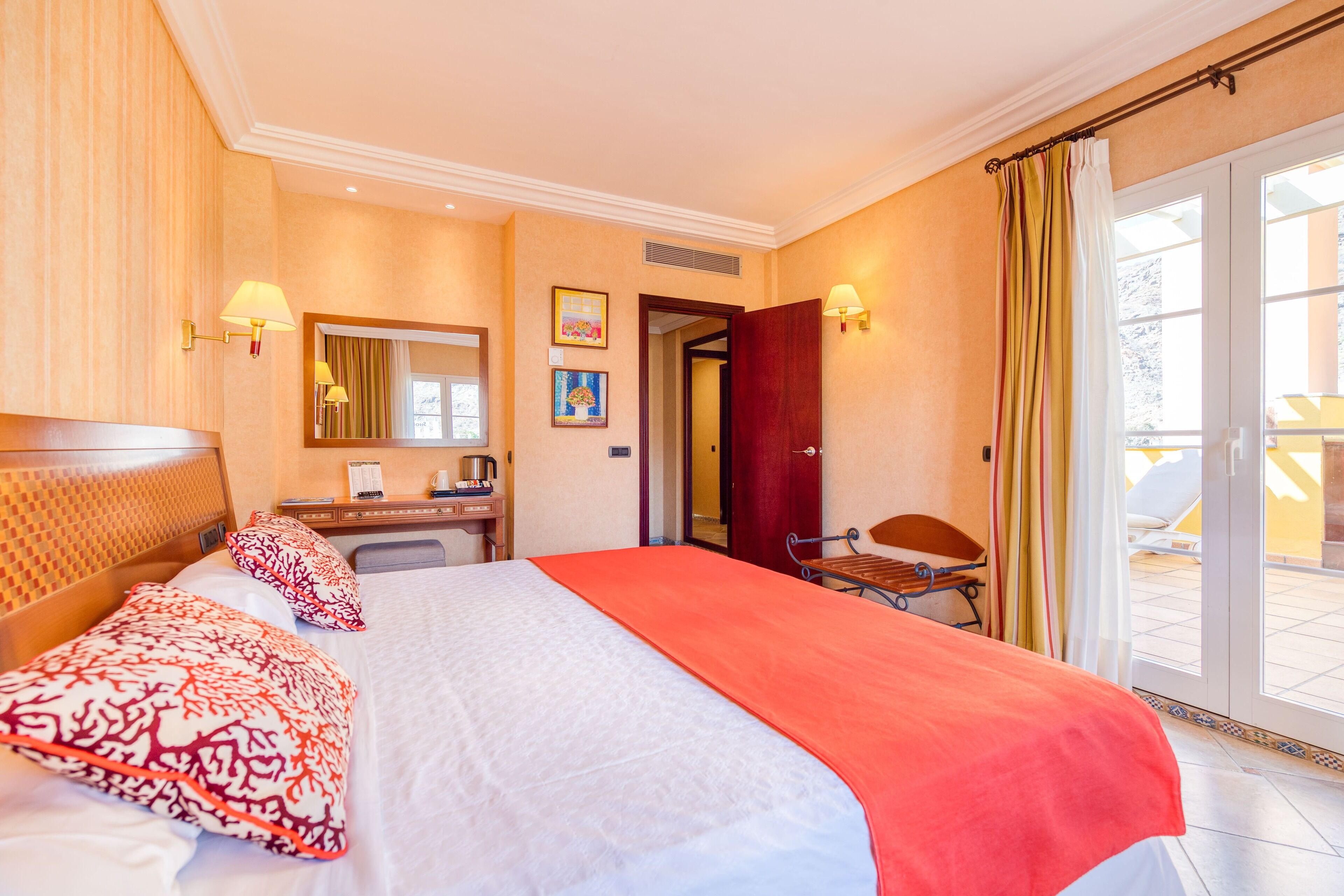 Une chambre d’hôtel avec un grand lit, un bureau, une chaise, un miroir et une porte-fenêtre coulissante donnant sur l’extérieur.