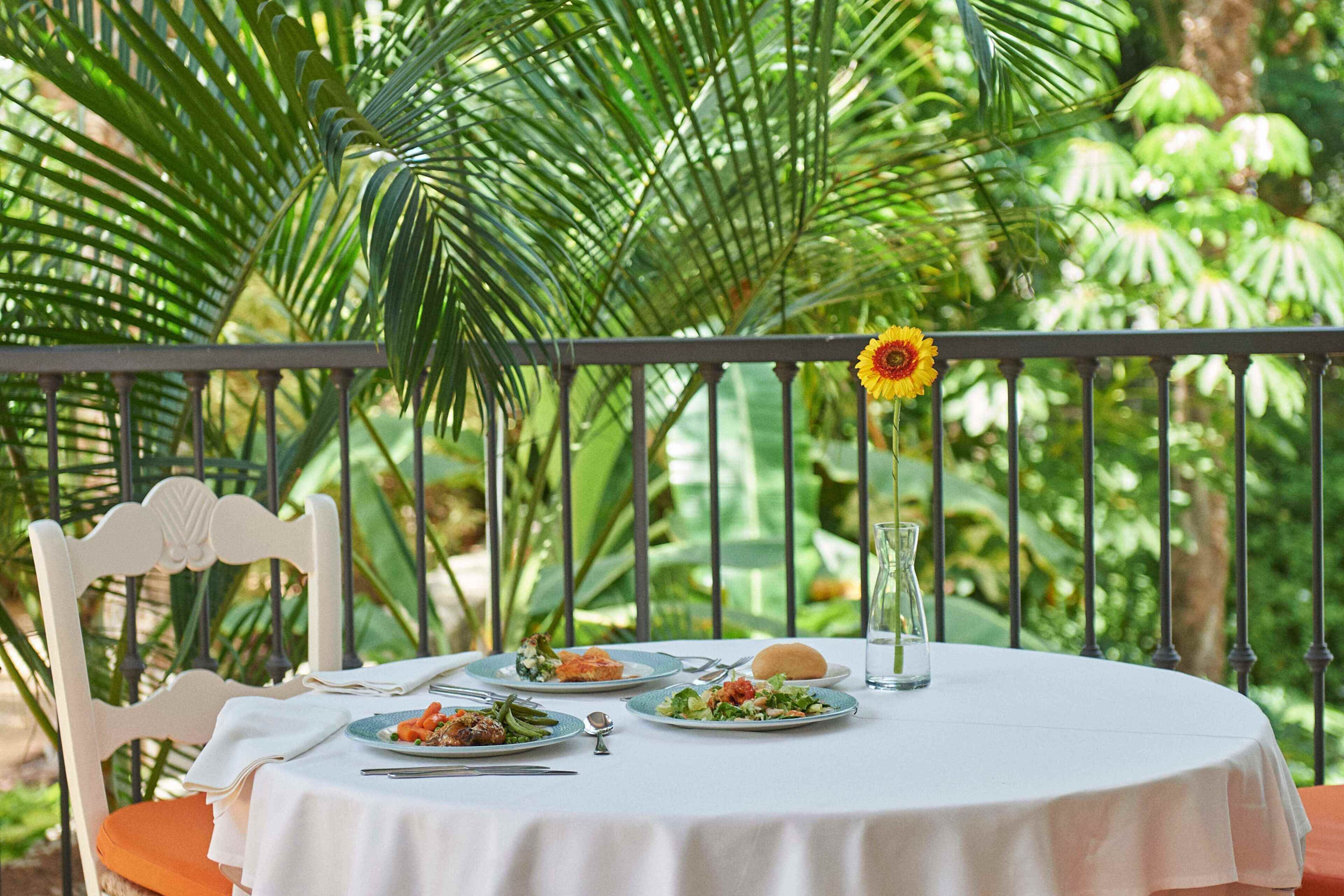 A round table with a white tablecloth, surrounded by chairs, set outdoors with a view of lush greenery and a metal railing.