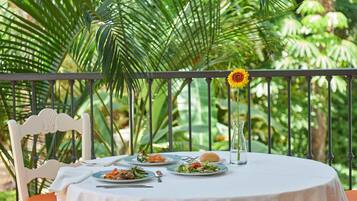 A round table with a white tablecloth, surrounded by chairs, set outdoors with a view of lush greenery and a metal railing.