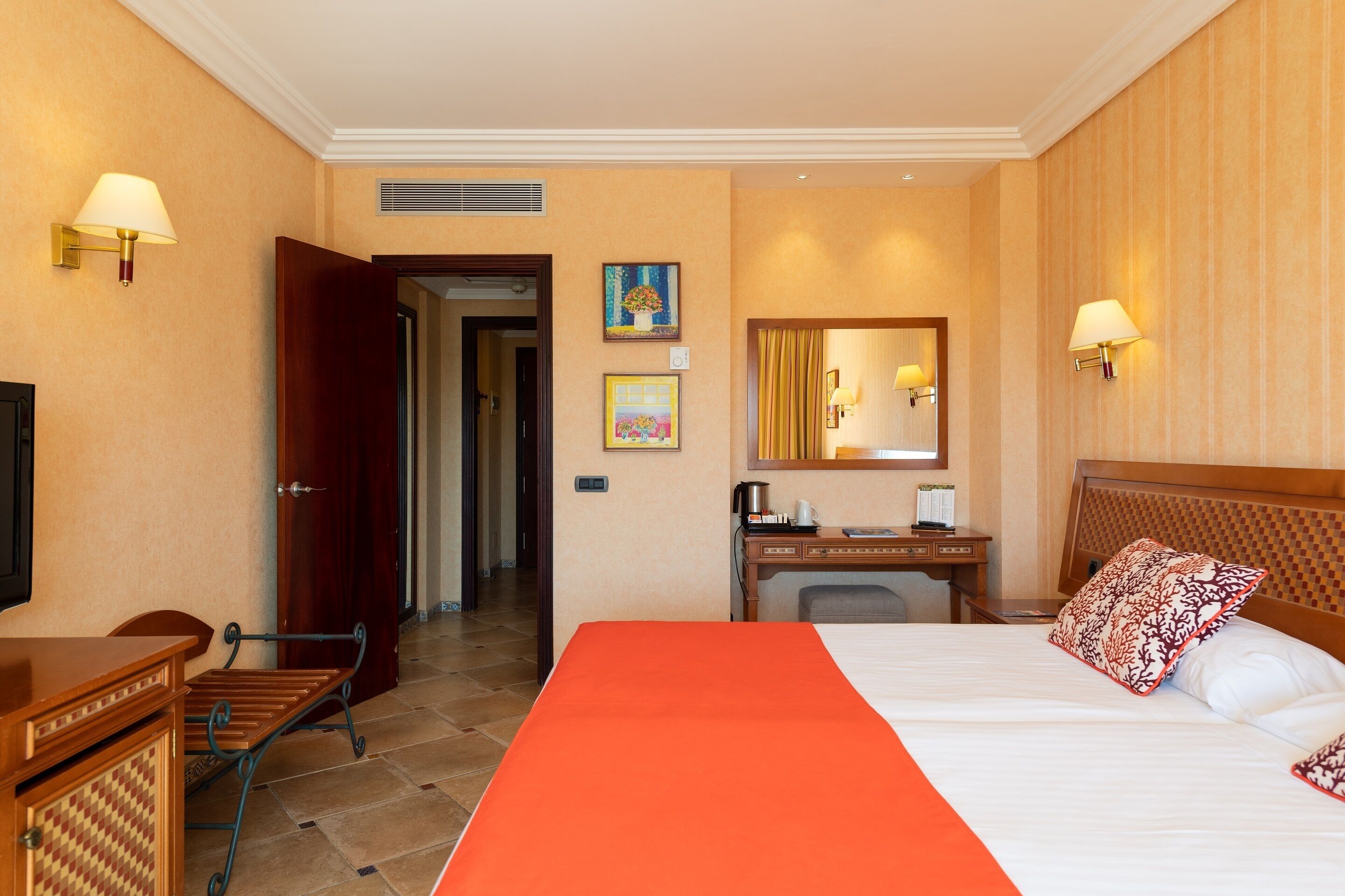A hotel room with a large bed, a desk, a chair, a TV, and a door leading to another room.
