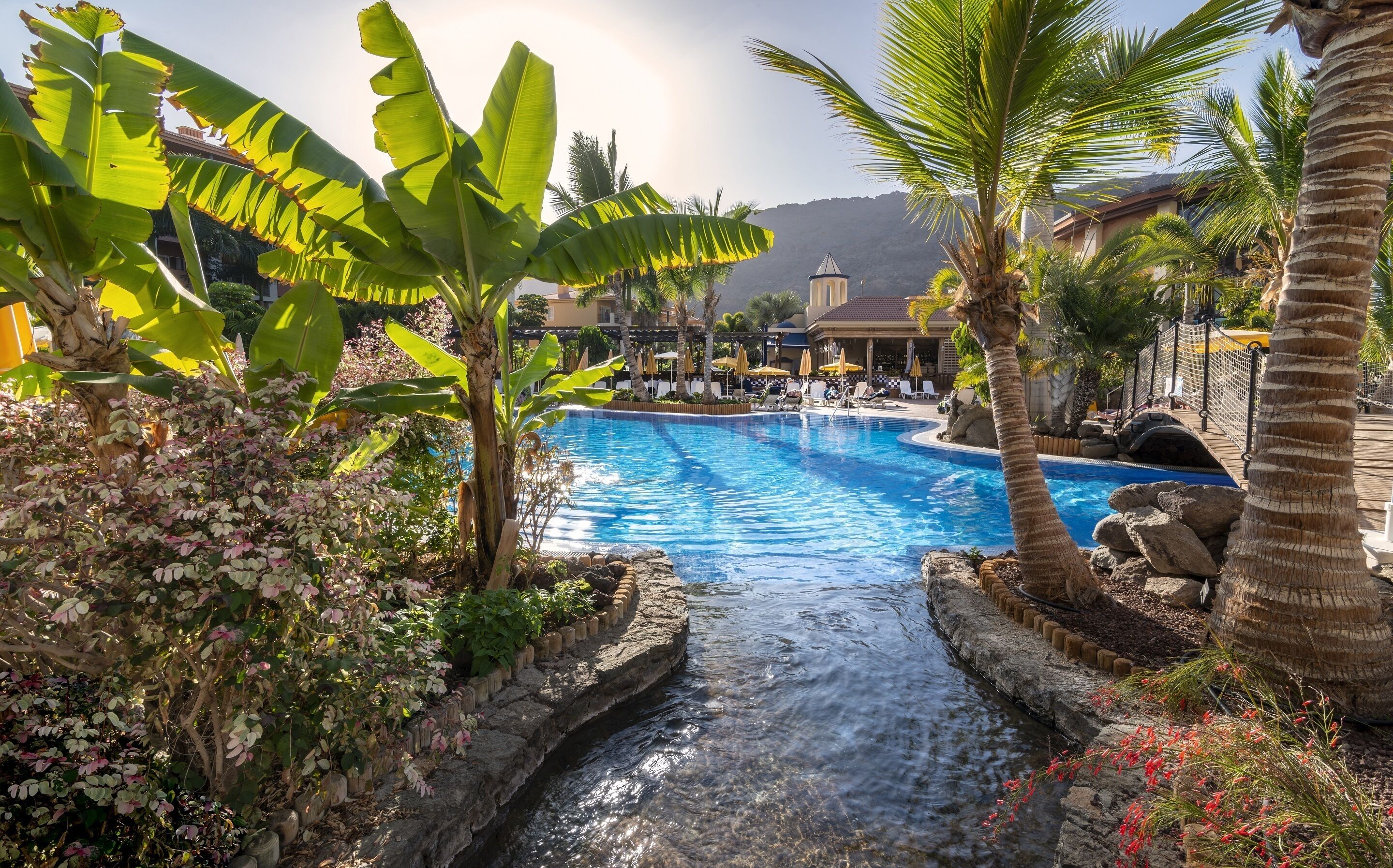 A pool surrounded by tropical plants and palm trees, with a view of a resort building in the background.