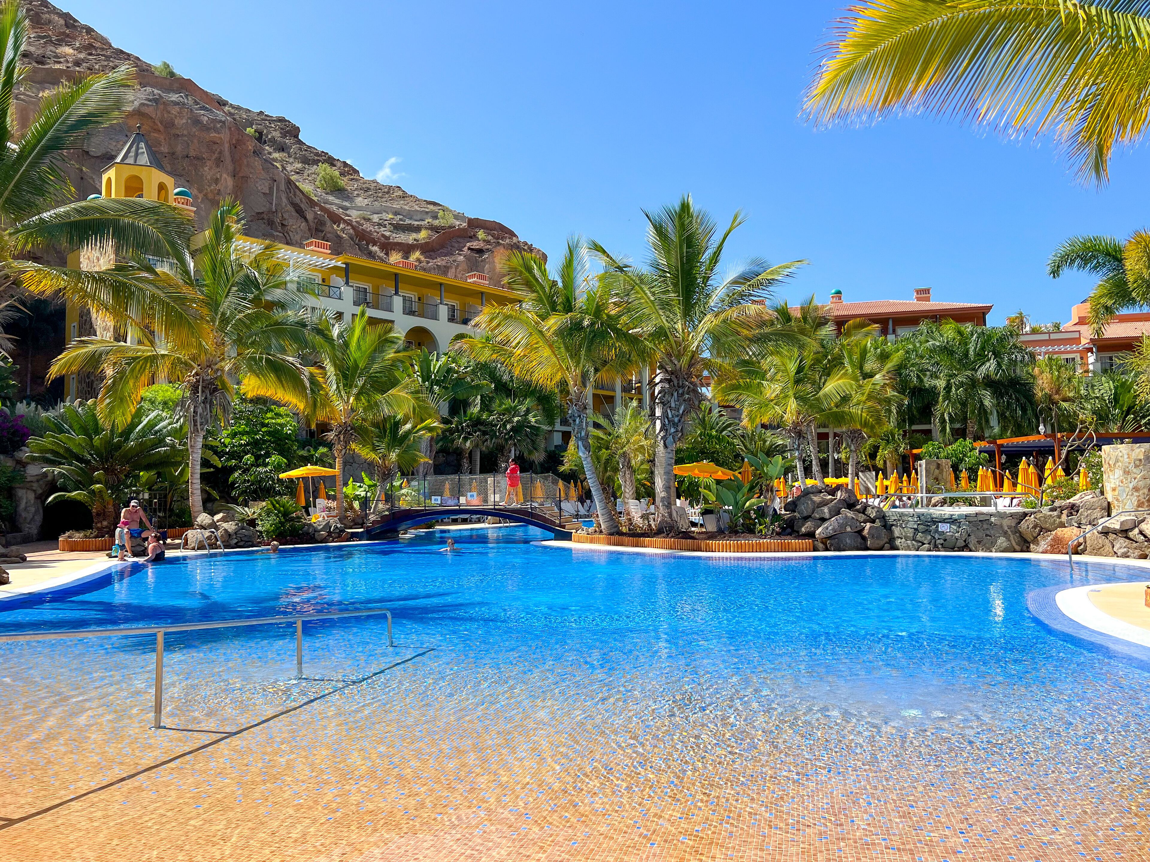Ein Resort-Pool mit Palmen, einer Brücke und einer Bergkulisse.