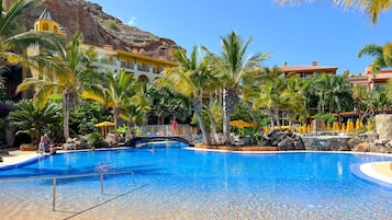 A resort pool with palm trees, a bridge, and a mountain backdrop.