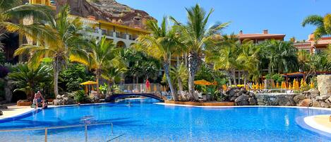 Ein Resort-Pool mit Palmen, einer Brücke und einer Bergkulisse.