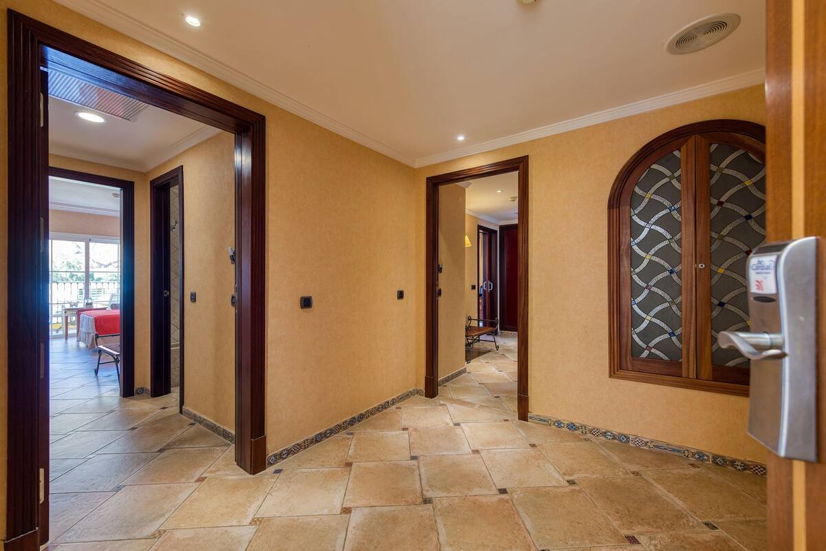 Un couloir doté d’un sol carrelé, de portes en bois et d’une vue sur une salle à manger où une nappe rouge est dressée.