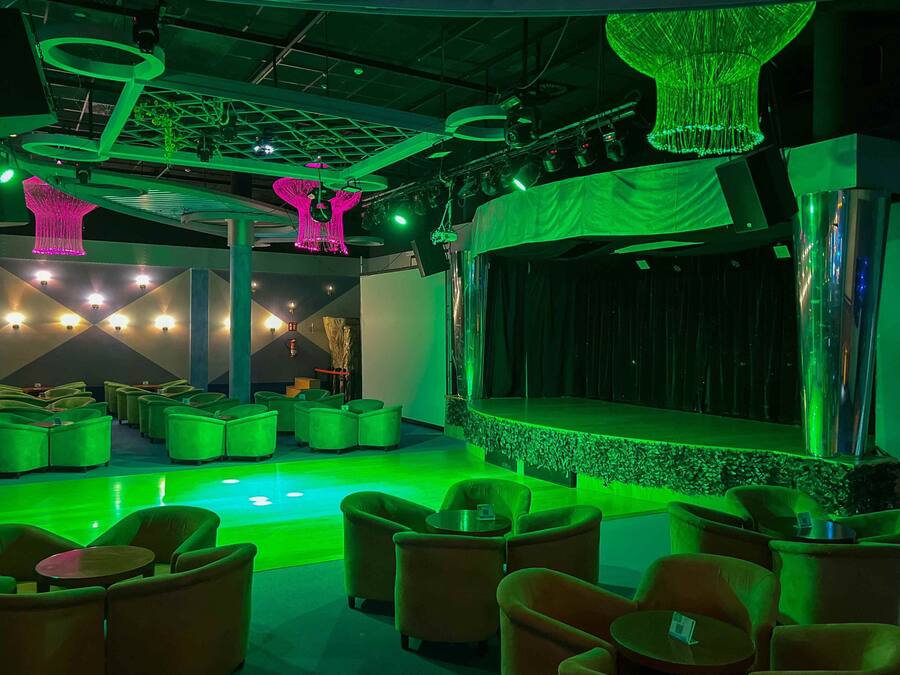 A lounge area with green ambient lighting, a stage, and multiple seating arrangements.
