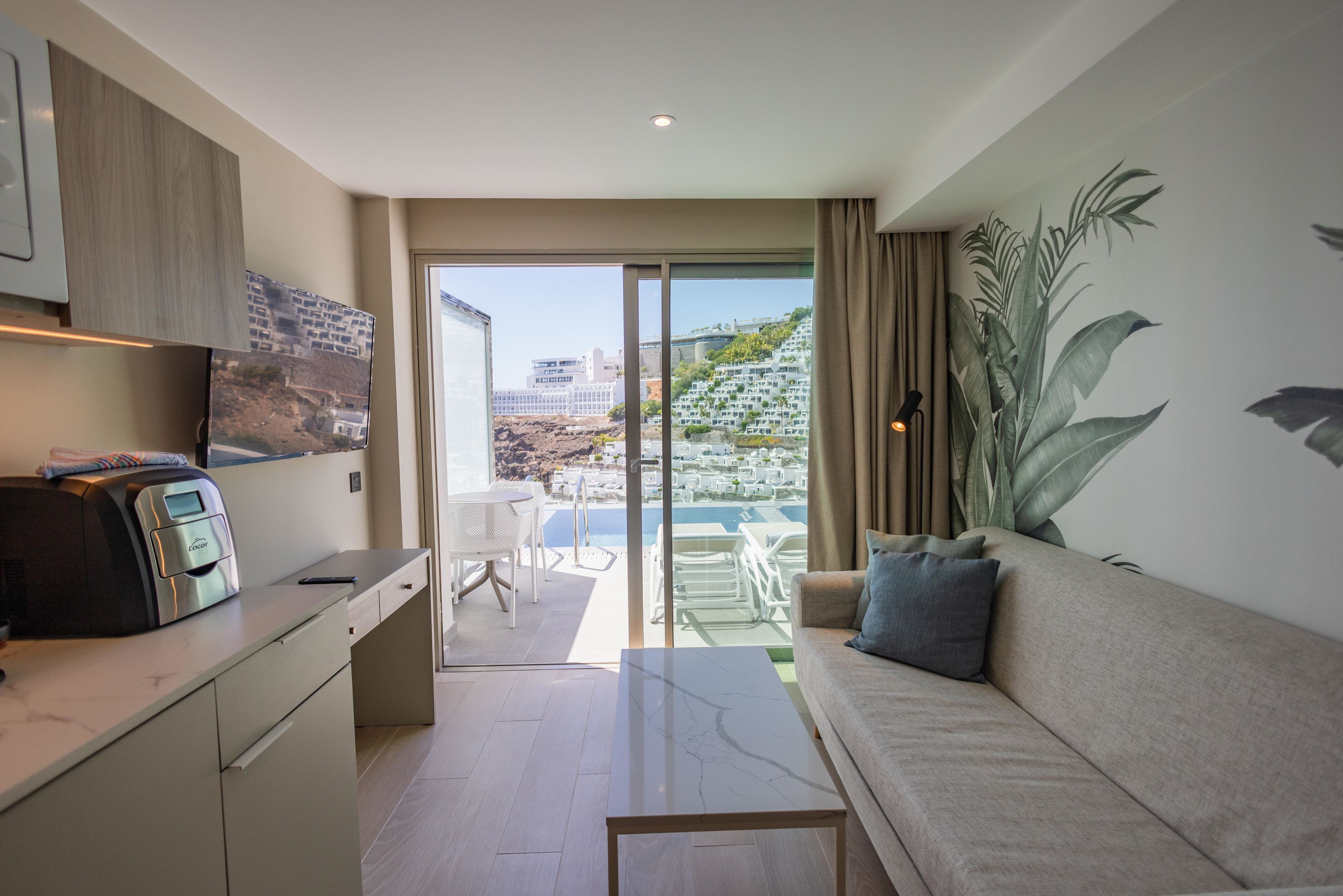 Una sala de estar moderna con un sofá, una mesa de centro y una zona de cocina con horno empotrado. Un amplio ventanal ofrece vistas a un paisaje urbano y a una piscina.