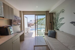 A modern living room with a sofa, a coffee table, and a kitchen area with a built-in oven. A large window offers a view of a cityscape and a pool. - Servatur Puerto Azul (Mogan)