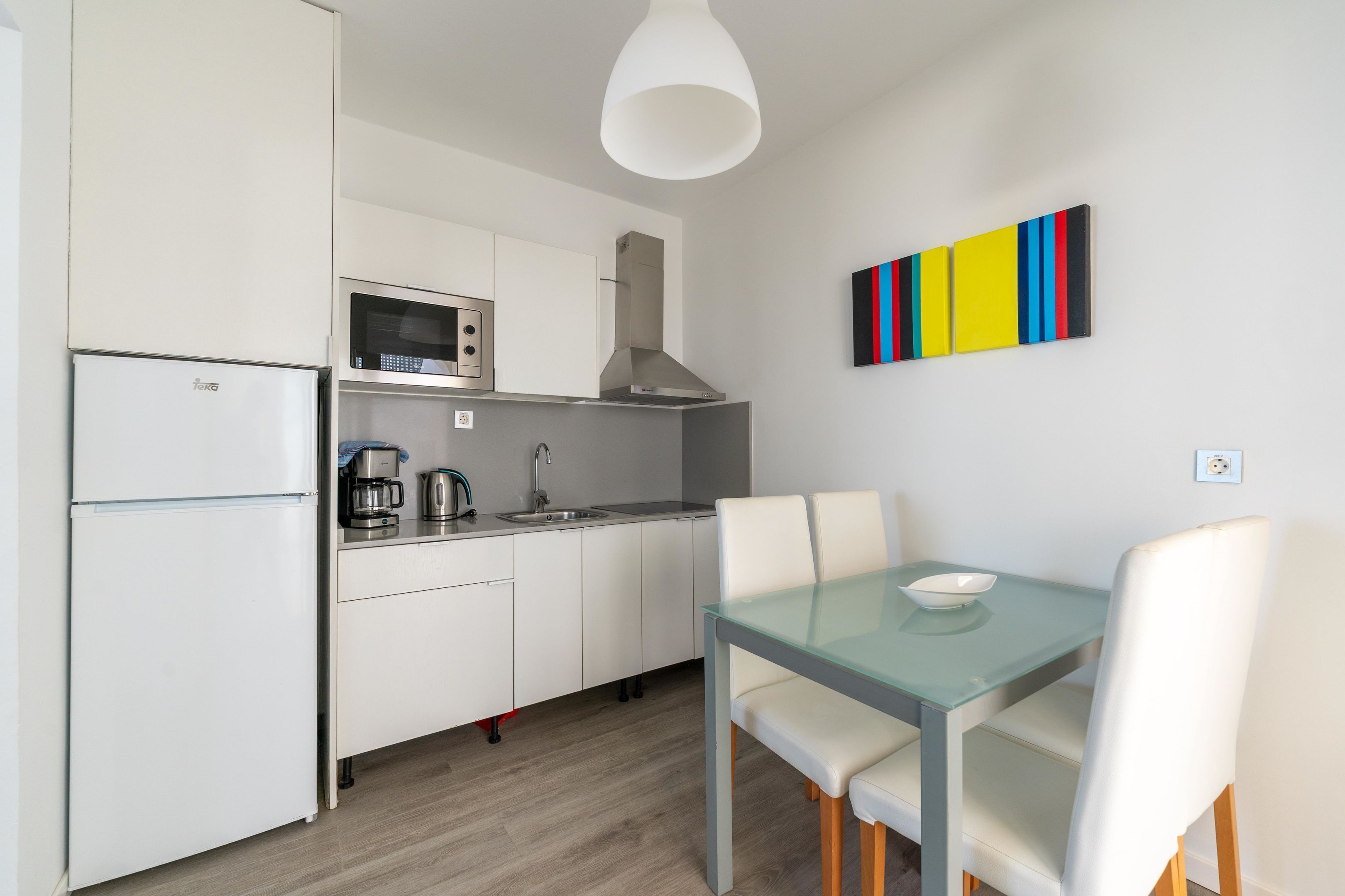 Una cocina moderna con armarios blancos, un microondas, una cafetera y un comedor con mesa de vidrio y sillas blancas.
