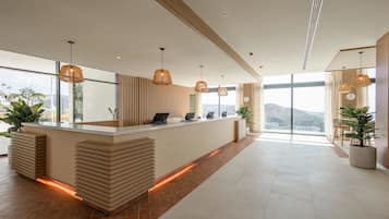 Un área de recepción moderna con un mostrador central, múltiples lámparas colgantes y grandes ventanales con vistas al exterior.