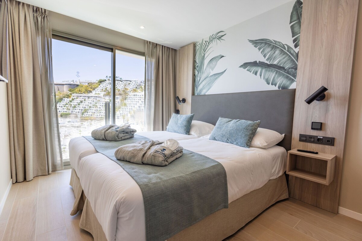 Una habitación de hotel moderna con una cama grande, vistas a la ciudad y un panel de control en la pared.