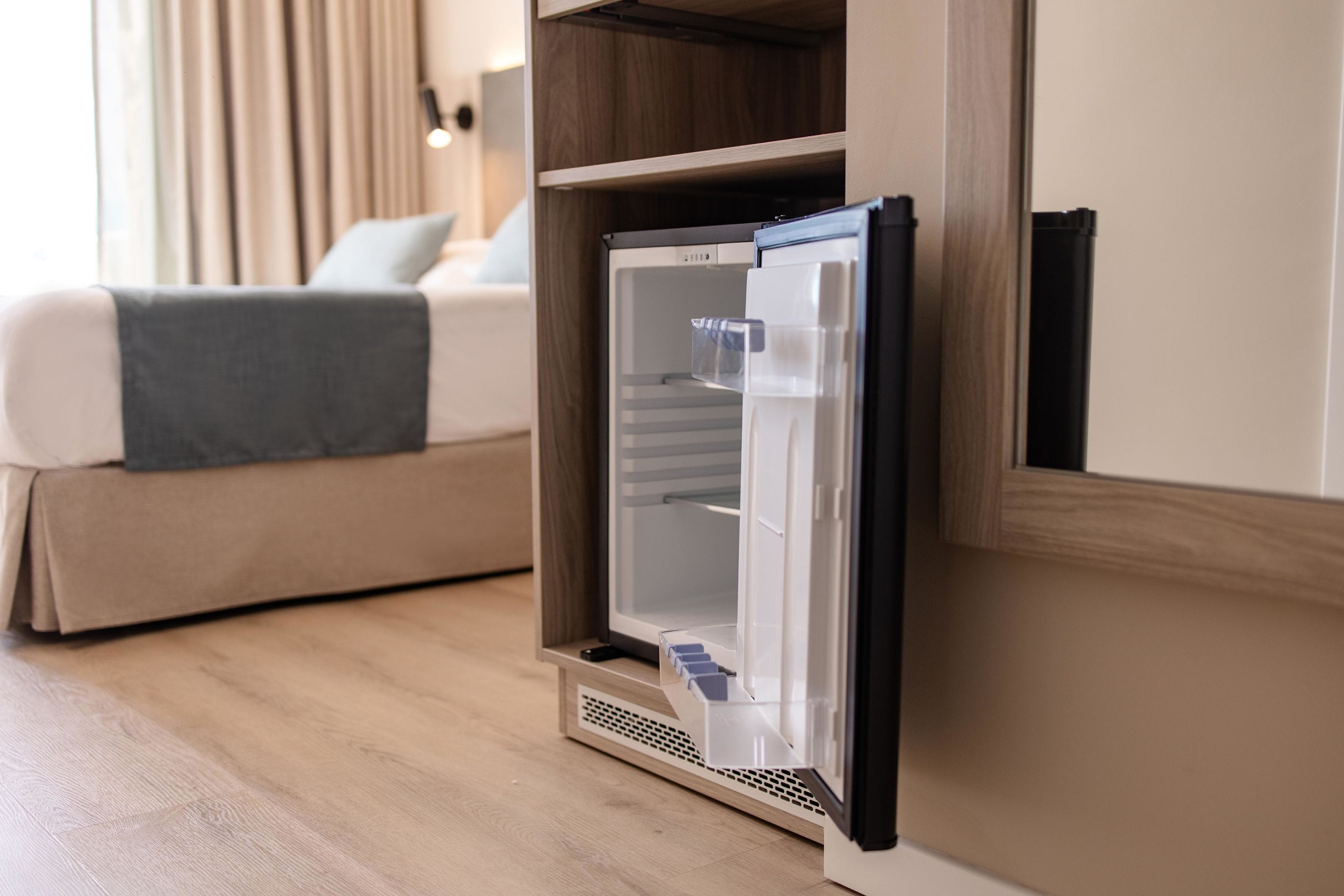 A compact refrigerator in a hotel room with a bed and curtains in the background.