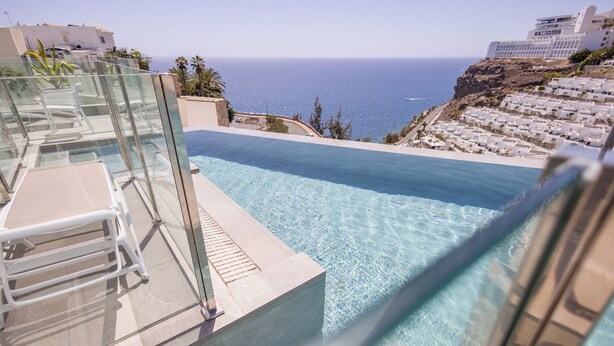 A clear blue infinity pool with a glass railing, overlooking a coastal area with buildings and a view of the sea. - Servatur Puerto Azul (Mogan)