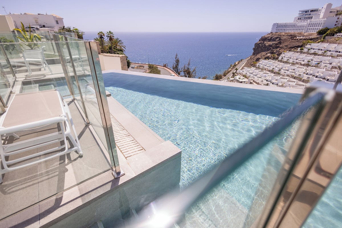 A clear blue infinity pool with a glass railing, overlooking a coastal area with buildings and a view of the sea.