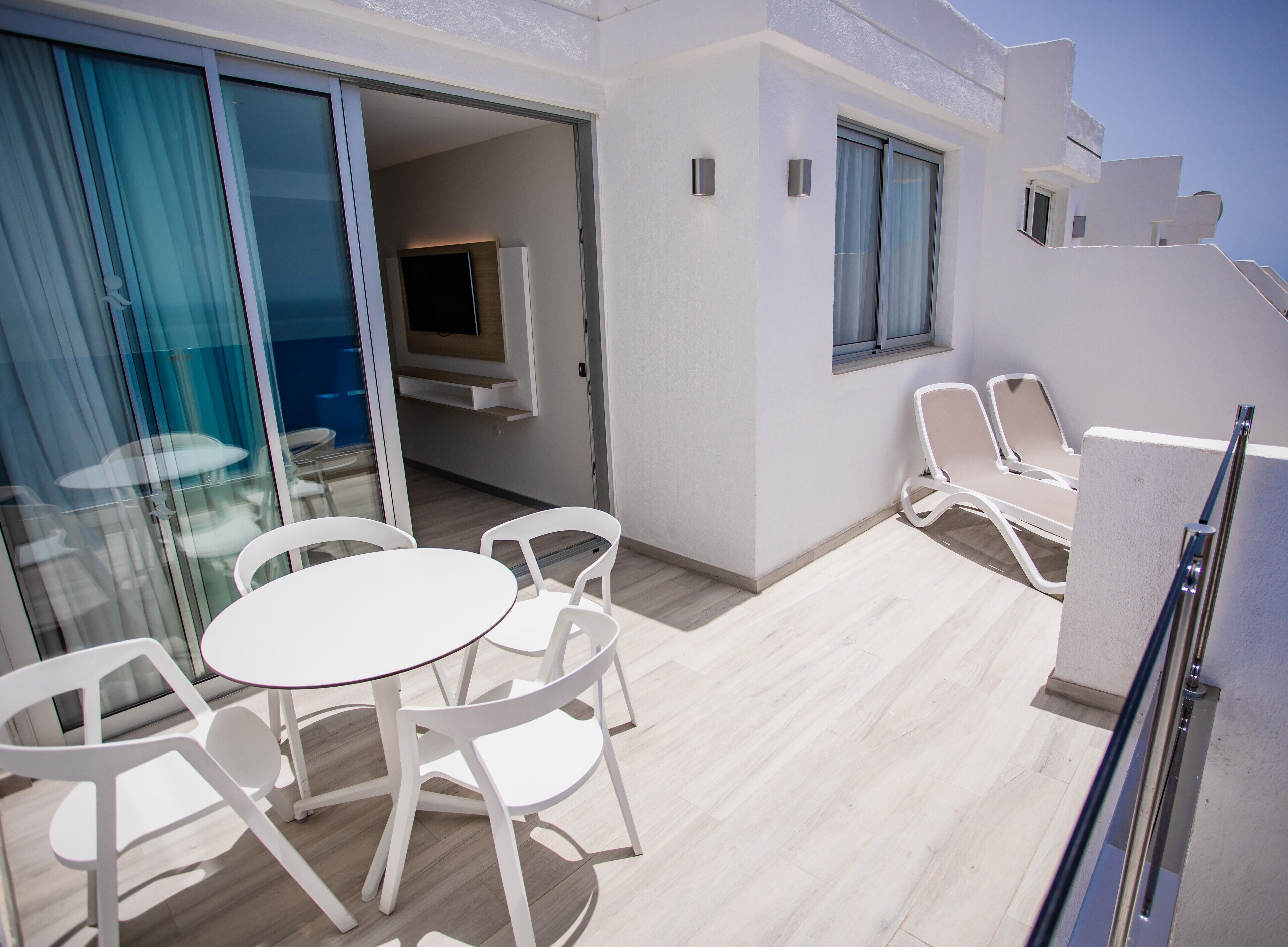 Varanda com cadeiras brancas e uma mesa, uma televisão fixada na parede e vista para o mar.