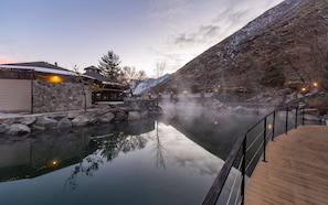 A serene hot spring area with a wooden deck, stone walls, and a building with outdoor seating.