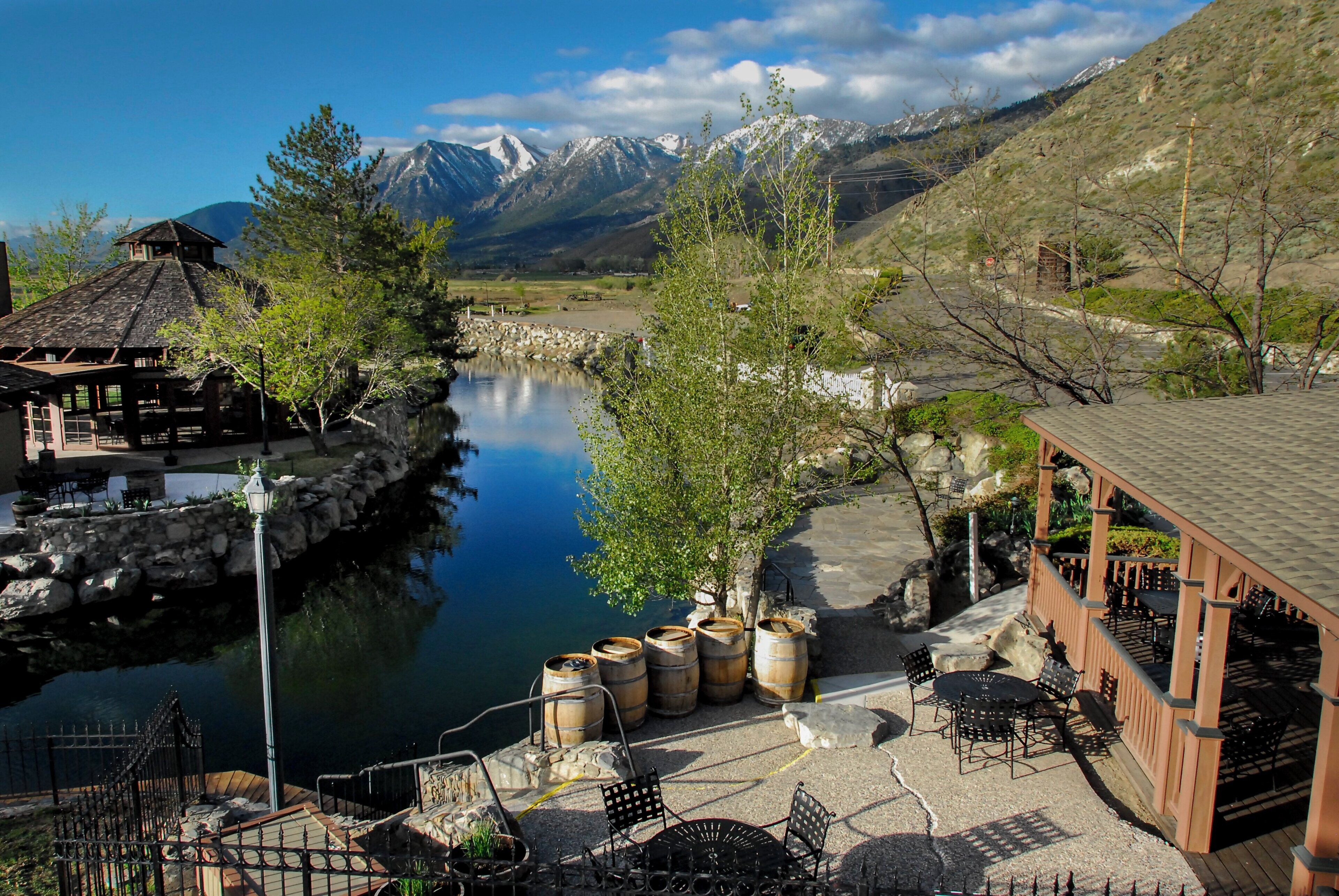 A riverside outdoor seating area with stone walls, wooden barrels, and a mountain backdrop.