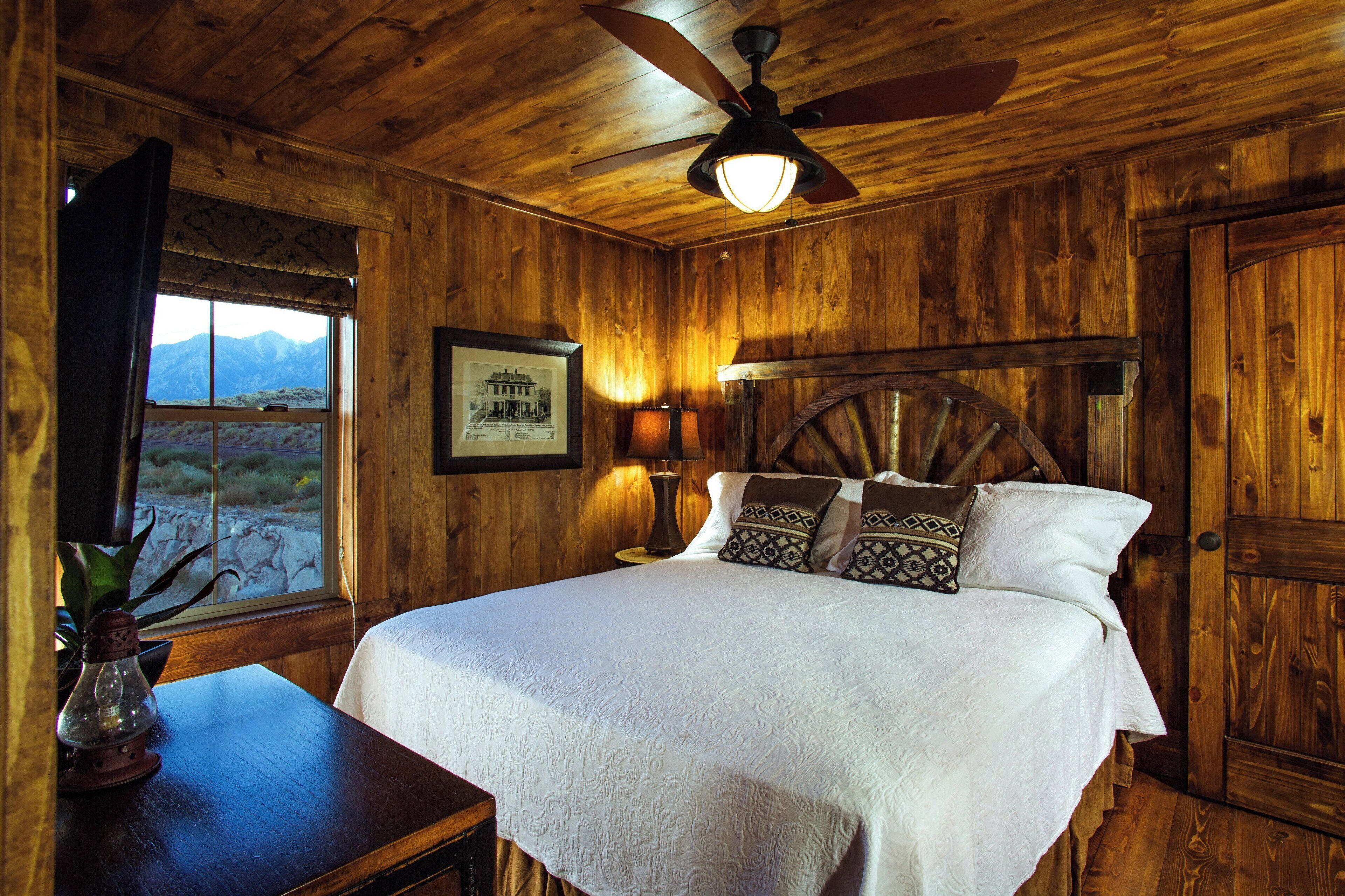 Una stanza in legno con un letto, una ventola a soffitto, una lampada, un quadro incorniciato e una finestra vista montagne.