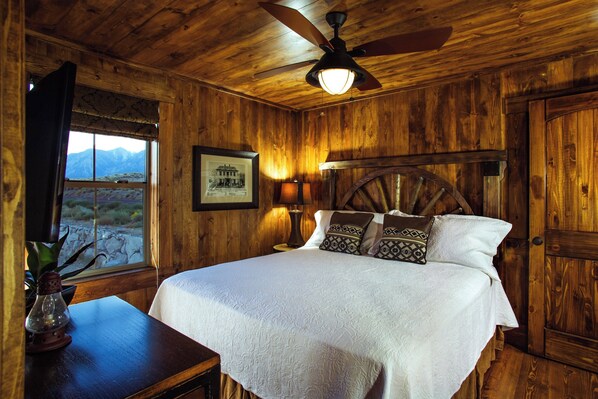 A wooden room with a bed, a ceiling fan, a lamp, a framed picture, and a window with a view of mountains.