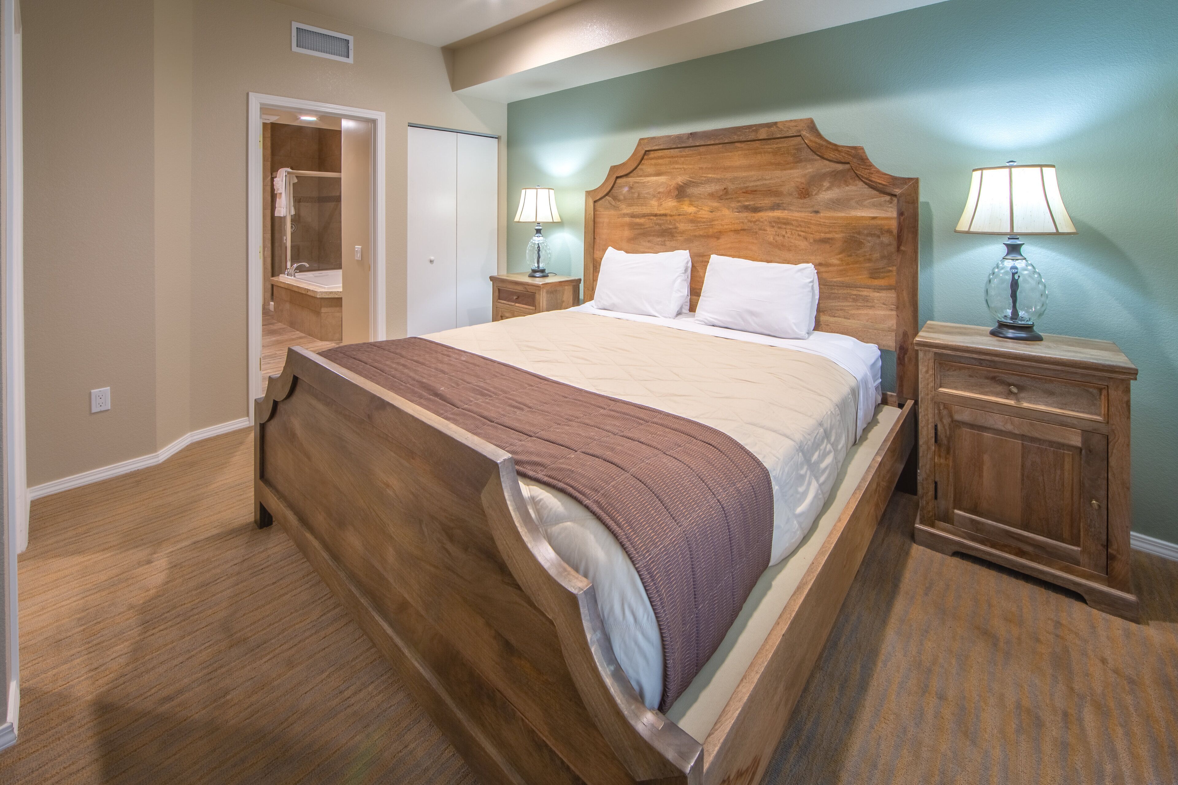 Un dormitorio con una cama de madera, dos mesitas de noche con lámparas, una mesita auxiliar de madera y una puerta que da a un baño.