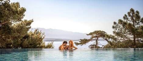 A couple in a clear infinity pool with a view of mountains and trees.