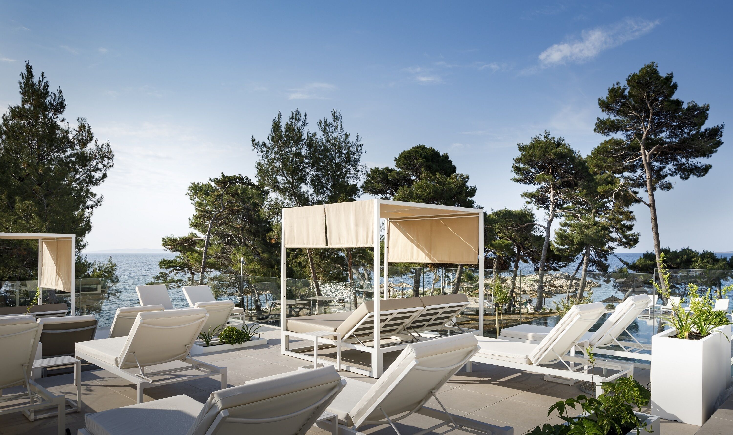 A poolside area with sun loungers and a canopy, overlooking a body of water with trees in the background.