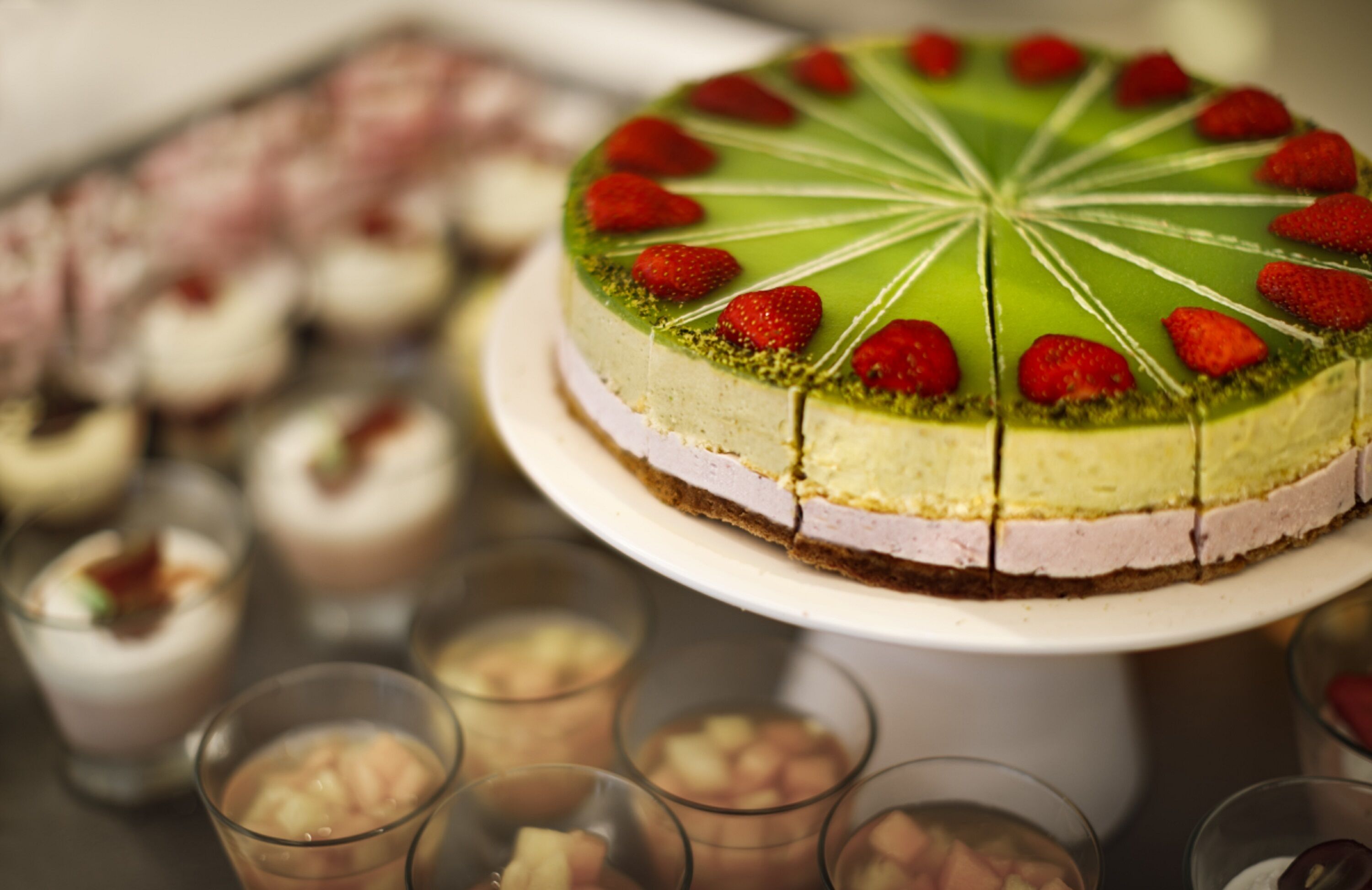A round cheesecake with strawberries on top, surrounded by various desserts and drinks.