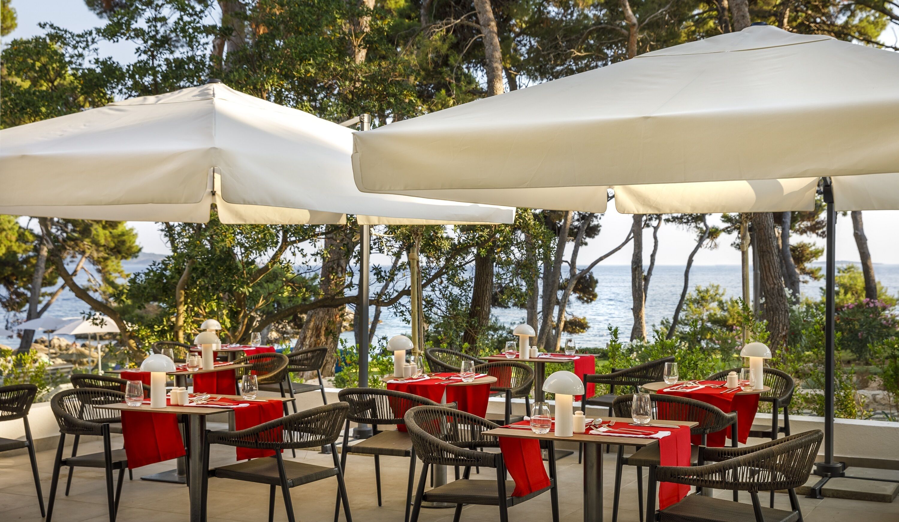 Een terras met tafels en stoelen, witte parasols en uitzicht op zee.