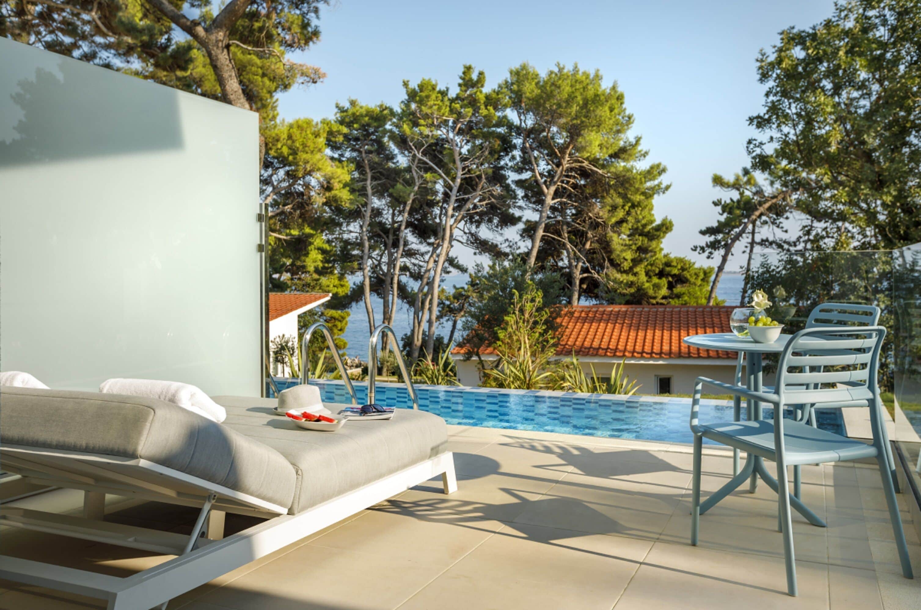 Ein Poolbereich mit Liegestuhl, Tisch und Stühlen sowie Blick auf das Meer und Bäume.
