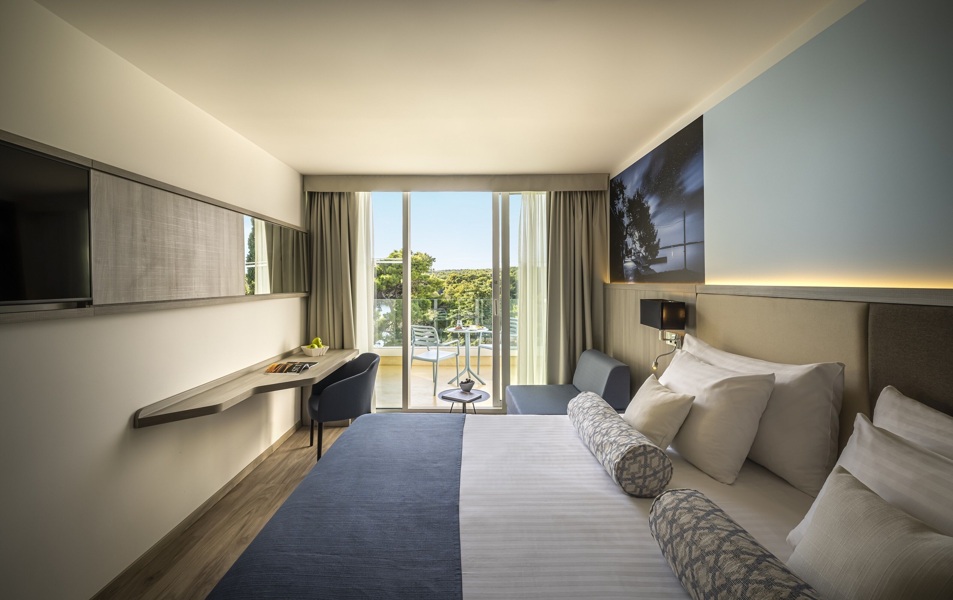 A hotel room with a bed, a desk, a chair, a TV, and a balcony with a view.