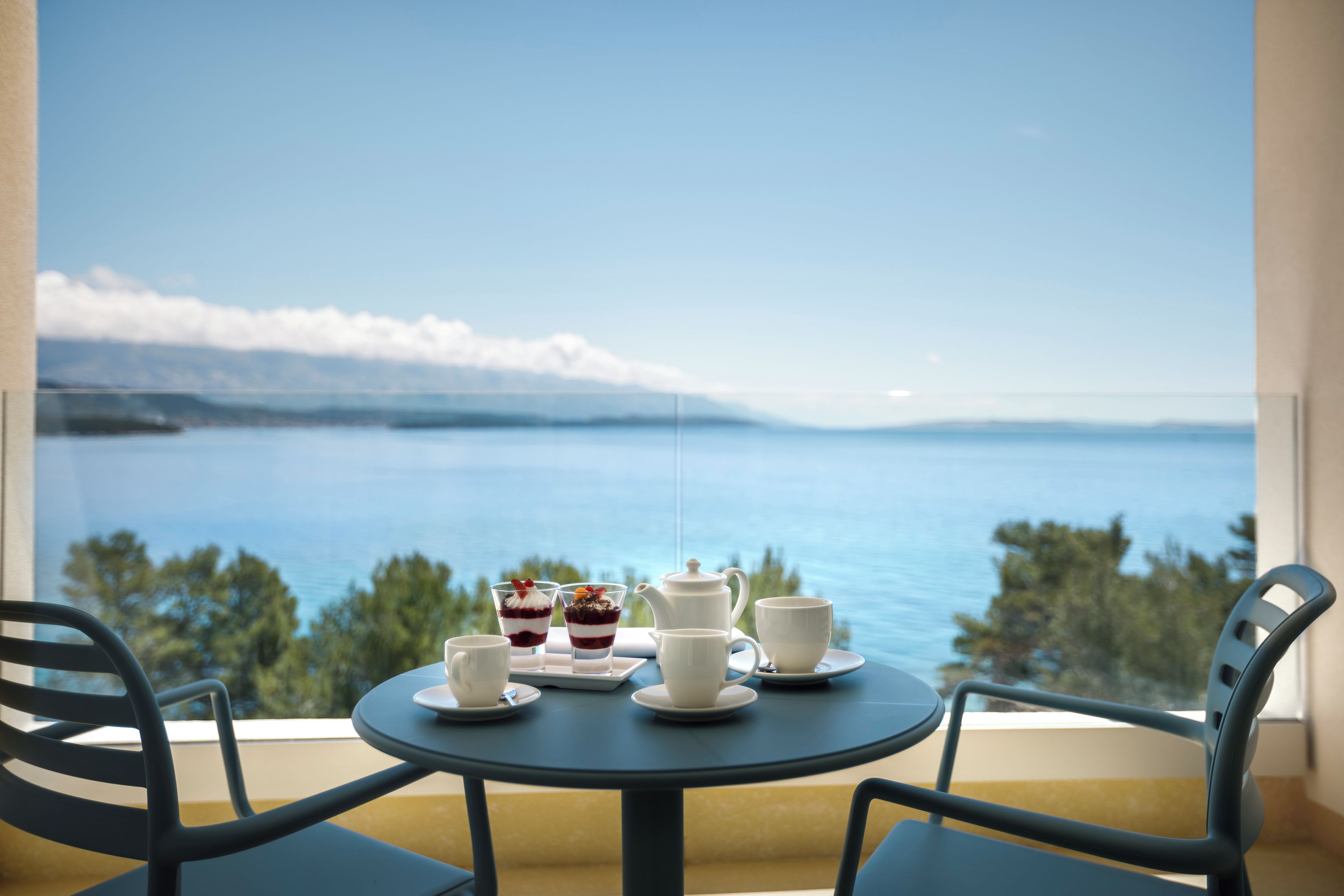 A table with two cups, a teapot, and a dessert on a balcony overlooking a body of water.