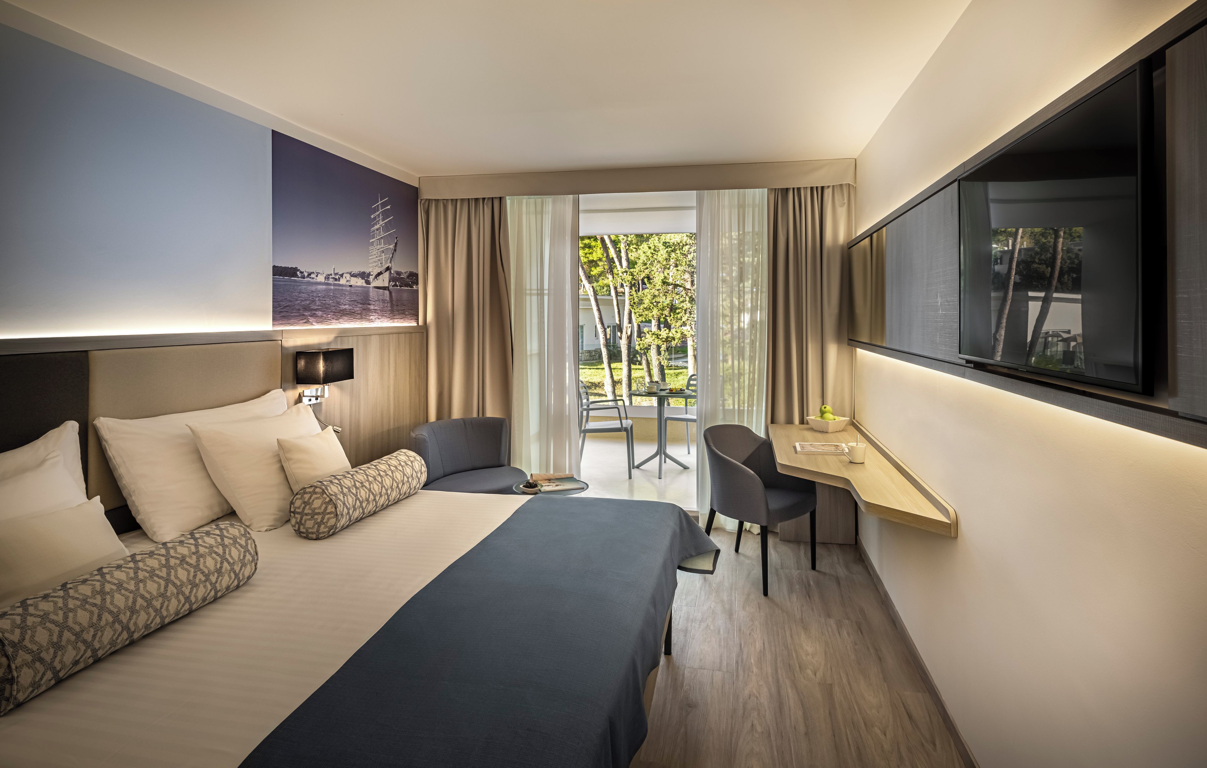 A hotel room with a bed, a desk, a chair, a TV, and a view of the outdoors.